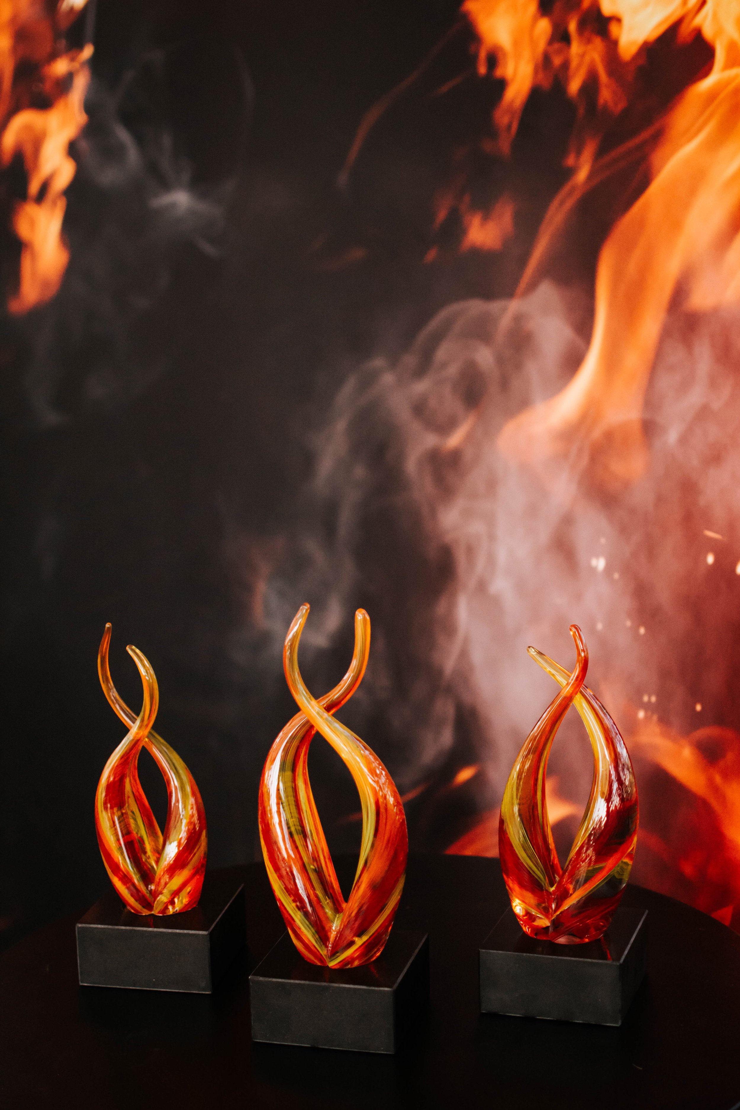 Four colorful glass sculptures with orange, yellow, and red flames are displayed on black stands, against a backdrop of a fiery scene with swirling flames and smoke.