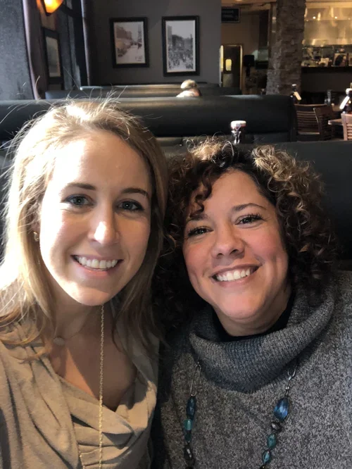 Two women smiling for a selfie in a restaurant or cafe with dark walls, framed pictures, and booth seating.