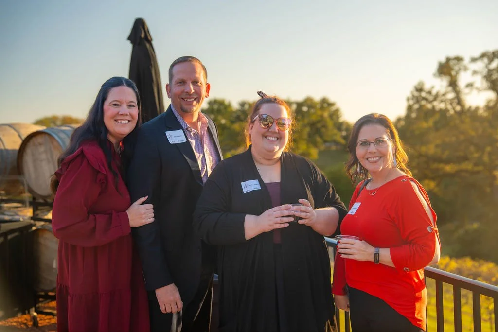 Four people smiling at an outdoor event during sunset, holding drinks, with trees and a balcony in the background.