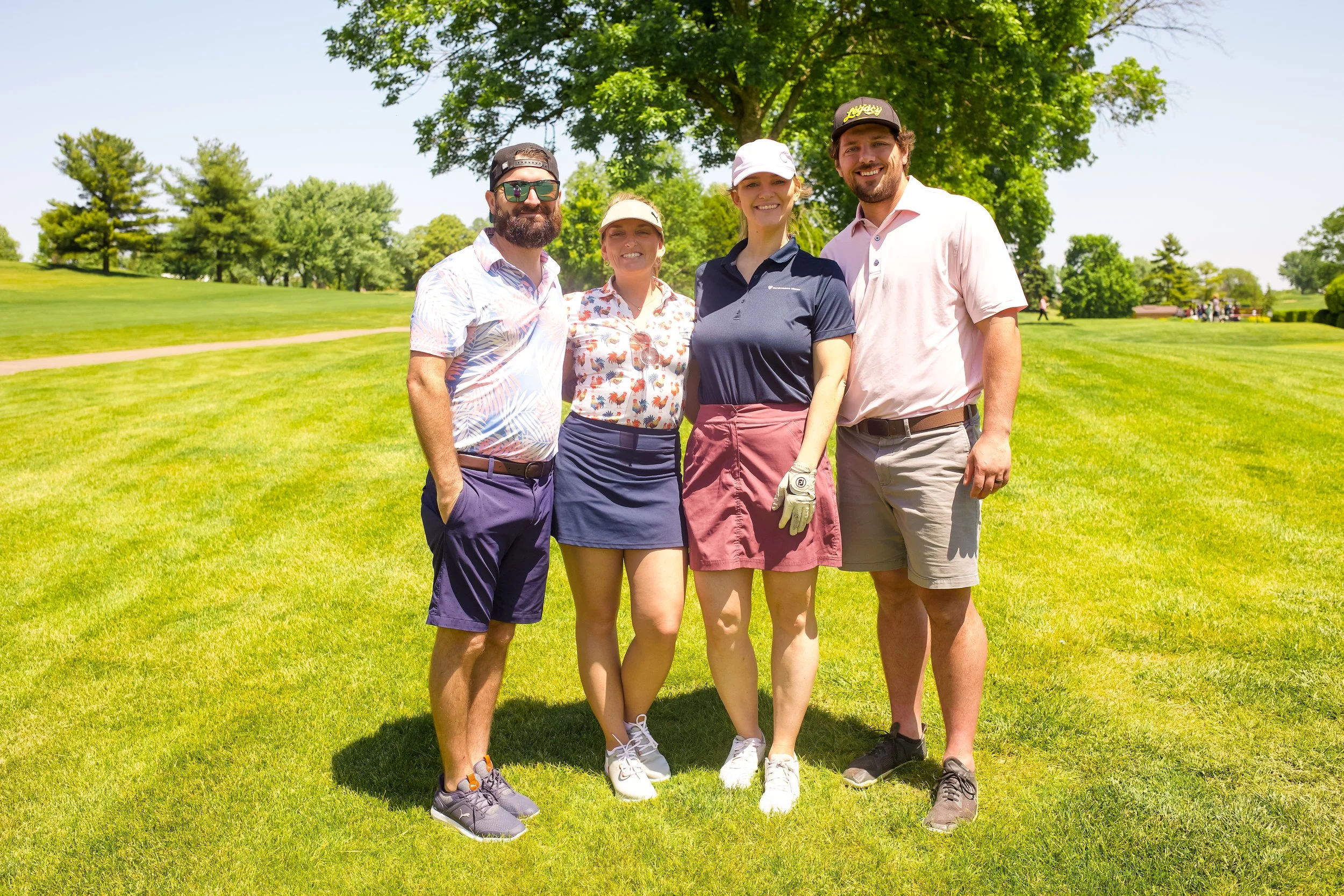 Four friends standing on a golf course, smiling, with trees and a clear blue sky in the background. They are wearing casual golf attire.