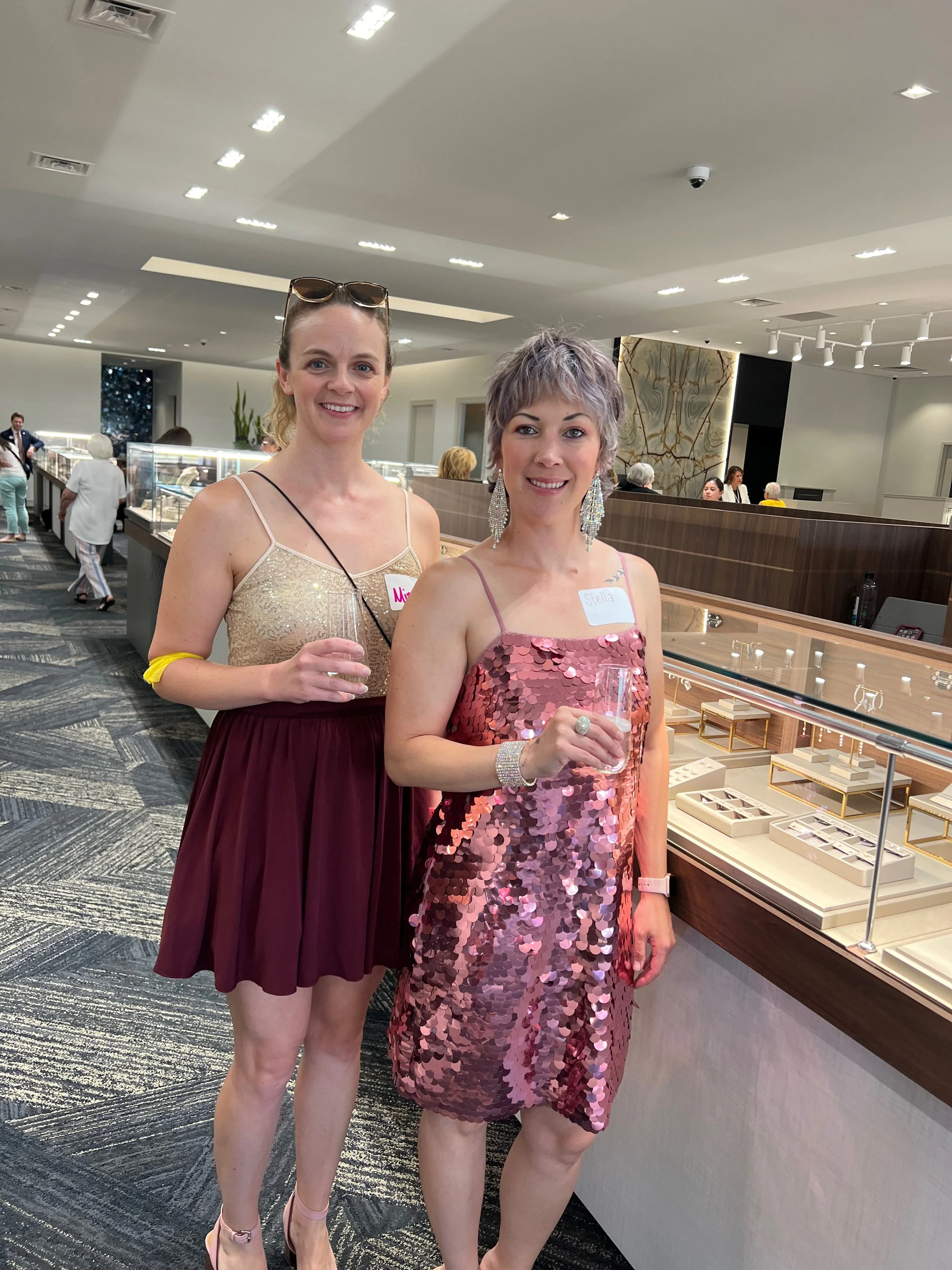 Two women in dresses at a gift shop or jewelry store, holding champagne glasses, smiling, with other shoppers in the background.