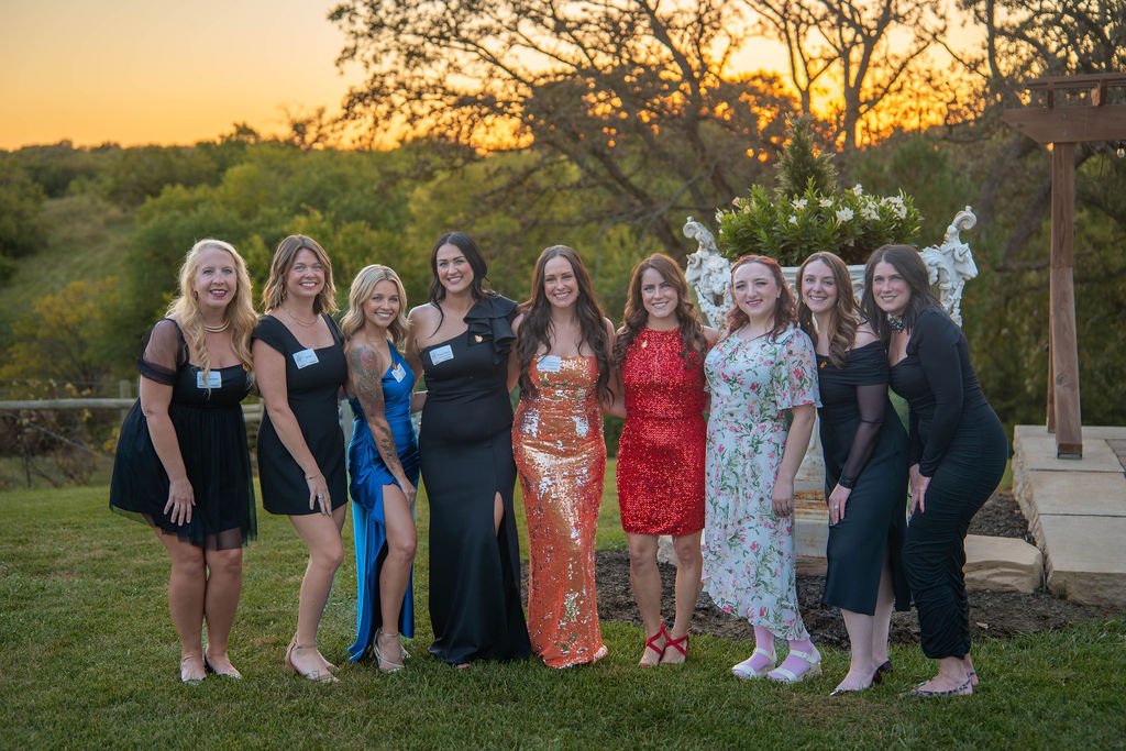 Group of nine women dressed in formal attire posing outdoors at sunset, with a decorative floral arrangement in the background.