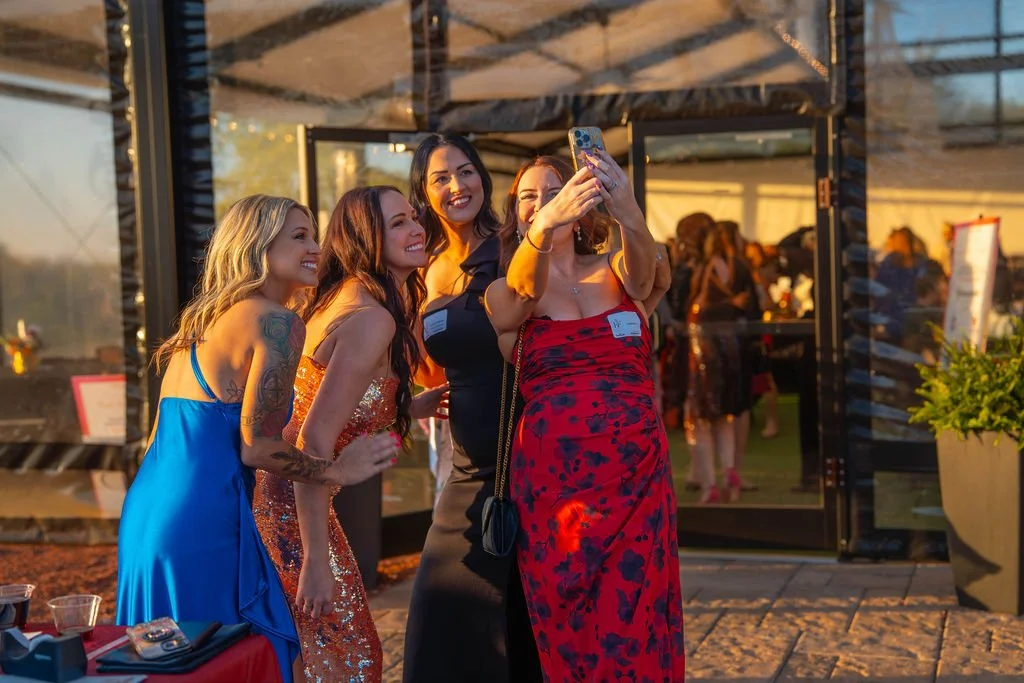 Four women taking a selfie outside a glass building at sunset, dressed in colorful dresses, smiling and posing for the photo.