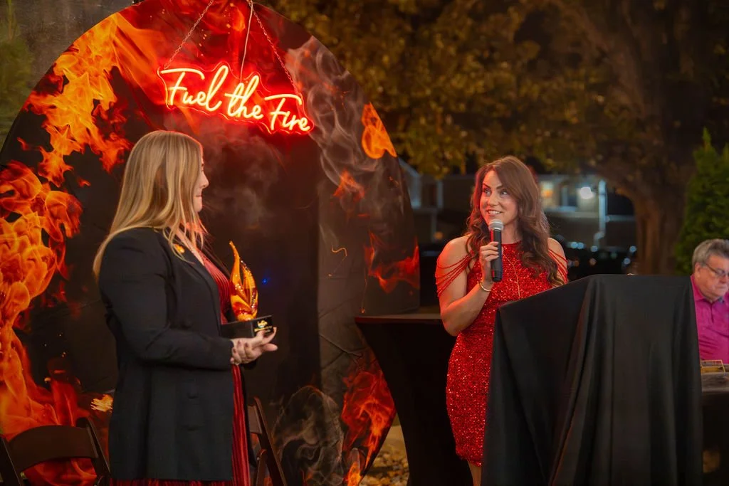 Two women at a fire-themed event, one is speaking into a microphone near a decorated backdrop with flames and the words "Fuel the Fire" written in neon. The woman on the left is holding an award.