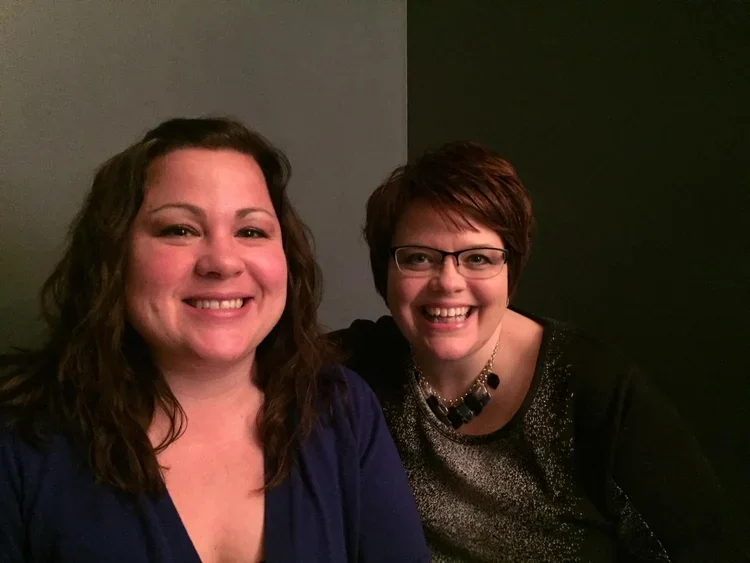 Two women smiling and sitting close together, one with long curly hair wearing a blue top and the other with short hair, glasses, and a black necklace, sitting in front of a two-toned wall painted gray and dark green.