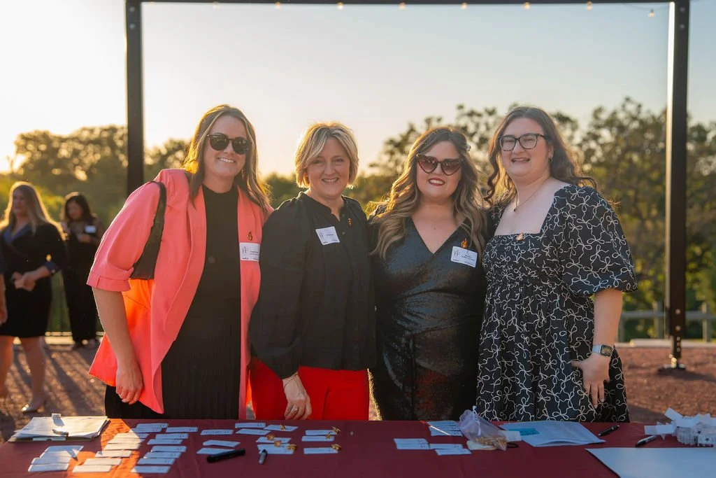 Four women standing outdoors at a registration table during sunset, smiling for the camera.