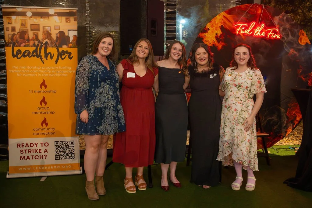 Five women standing together at a professional event, smiling for the camera, with a colorful background and a large sign about women leadership and mentorship to their left.