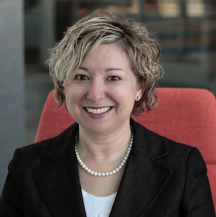 A smiling woman with short, wavy blonde hair, wearing a black blazer, a pearl necklace, and small earrings, sitting on an orange chair.