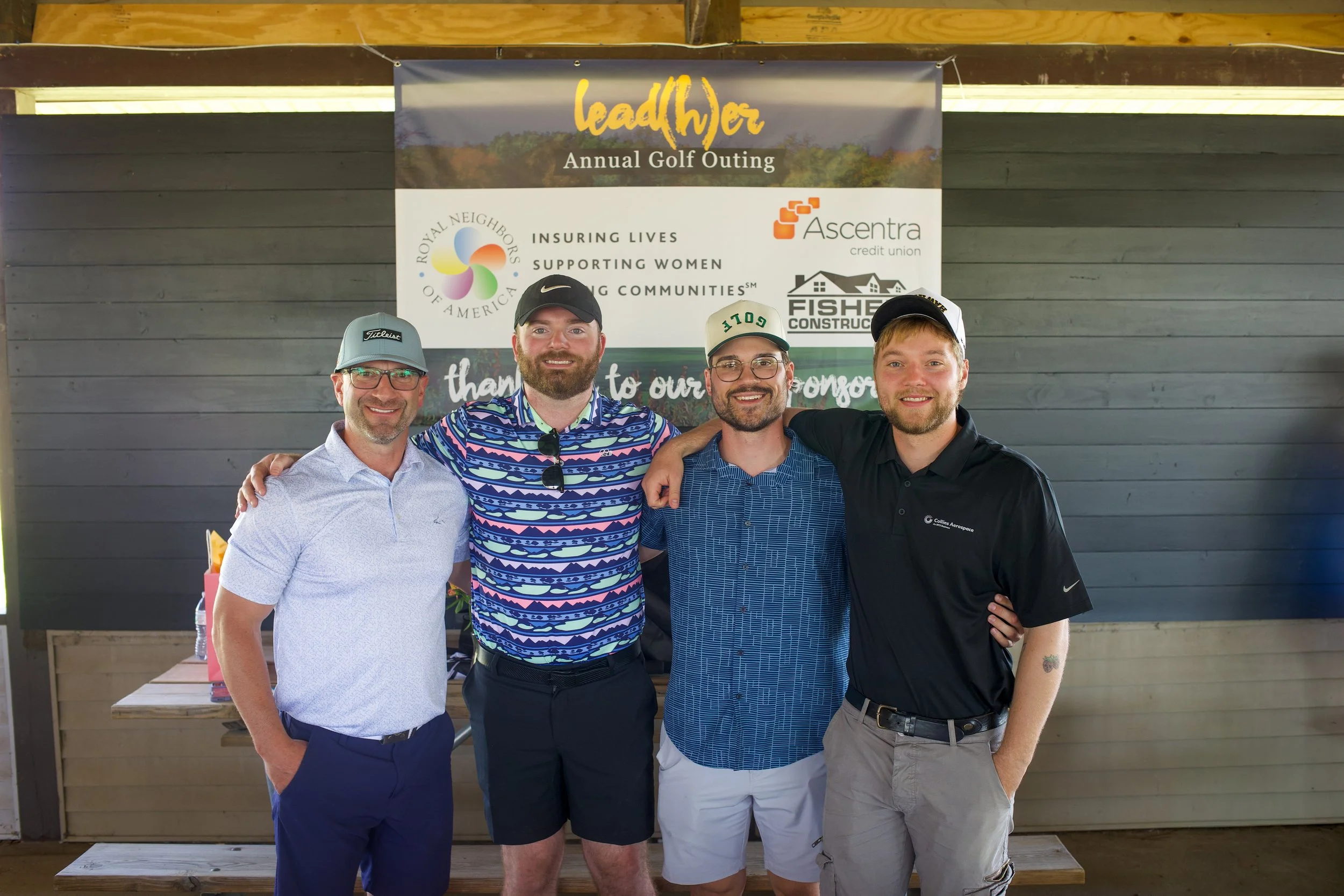 Four men standing together in front of a banner at an outdoor event, smiling for the photo. The banner behind them reads 'lead(h)er Annual Golf Outing' and features sponsor logos, including Royal Neighbors of America, Ascentra Credit Union, and Fishe