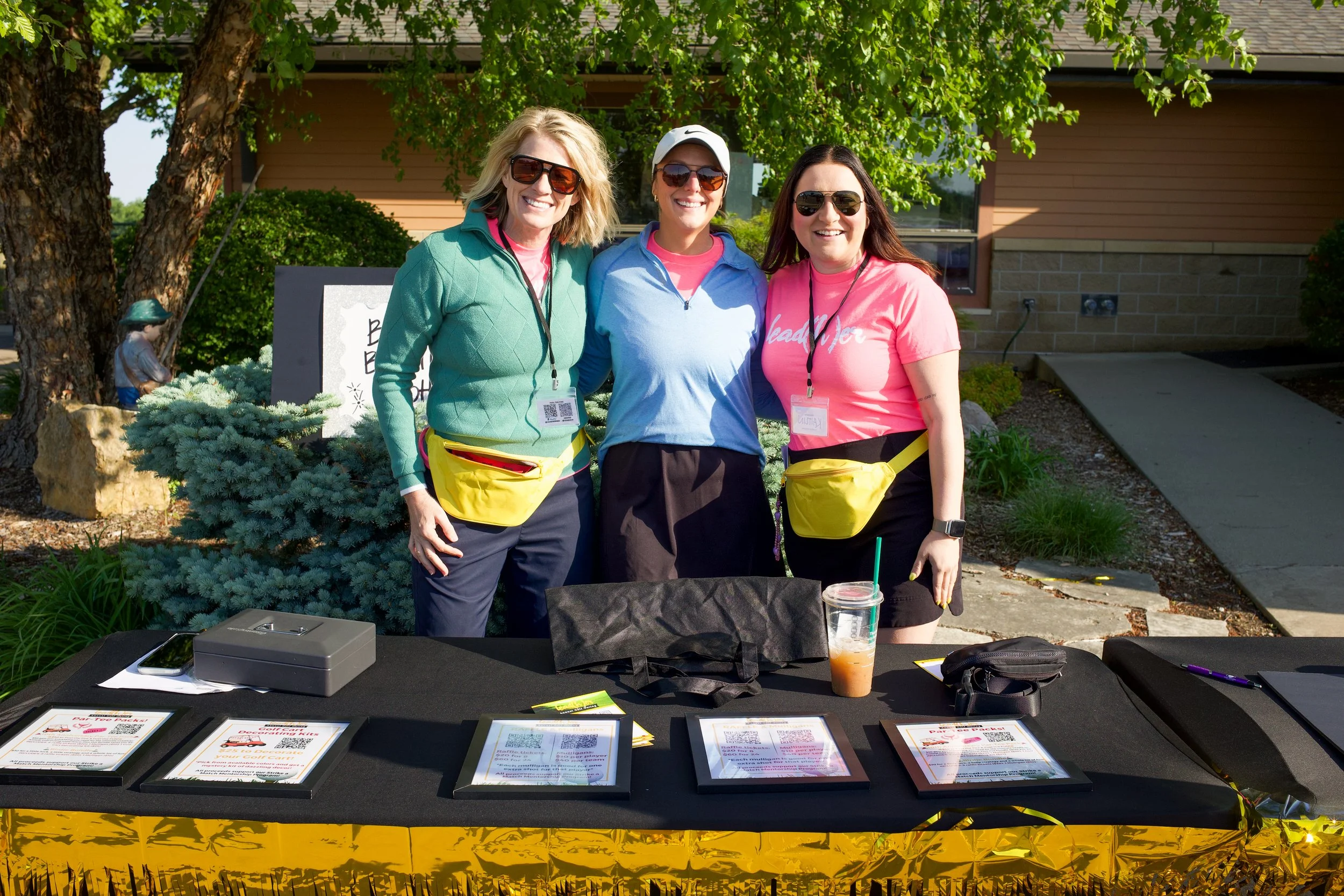 Three women standing outdoors behind a table with signs and flyers, smiling and wearing casual clothes with sunglasses, in front of a building and green trees.