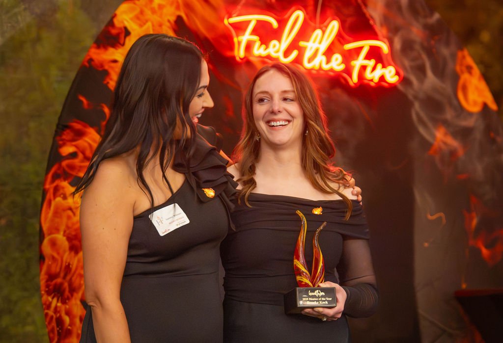 Two women in black dresses celebrating at an event. One is presenting, and the other is holding an award sculpture. Behind them is a fiery backdrop with a neon sign that reads "Fuel the Fire."