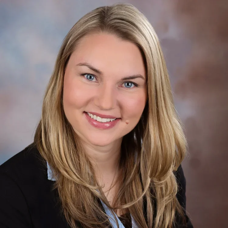 A woman with long blonde hair, blue eyes, and a friendly smile posing for a professional headshot against a blurred background.