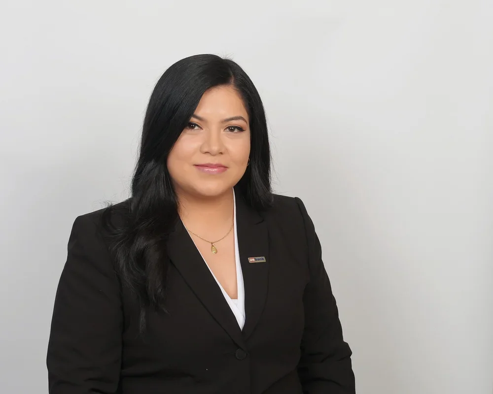 A woman with long black hair wearing a black suit, white blouse, and a gold necklace, standing against a plain light gray background.