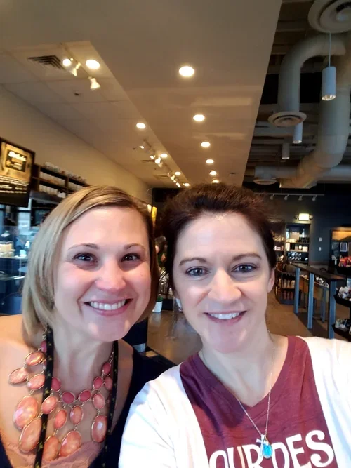 Two women smiling for a selfie inside a well-lit store or cafe, with shelves and ceiling fixtures visible in the background.