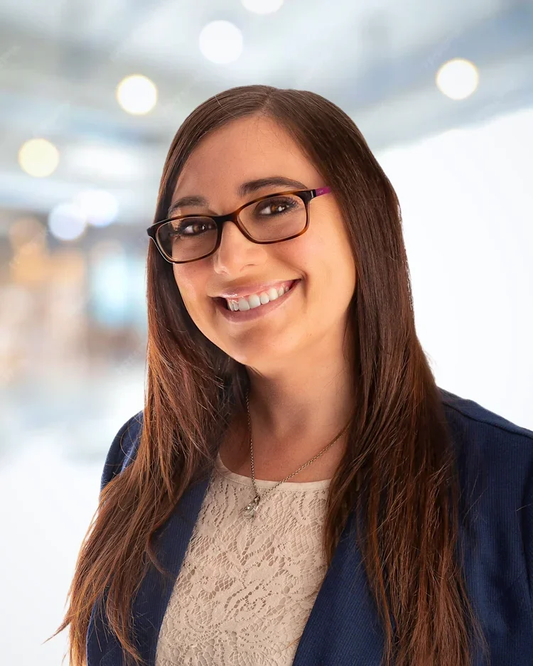A smiling woman with long brown hair, wearing glasses, a cream lace top, and a navy blazer, standing indoors with blurred lights in the background.
