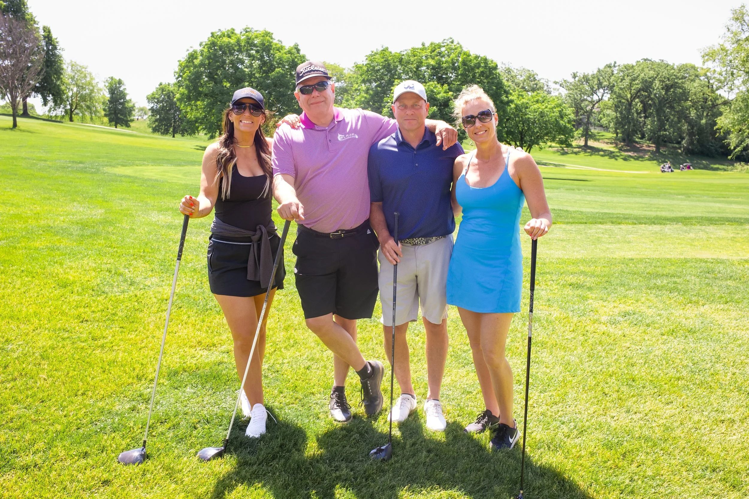 A group of four people smiling and posing with golf clubs on a golf course, surrounded by green grass and trees under a bright, sunny sky.