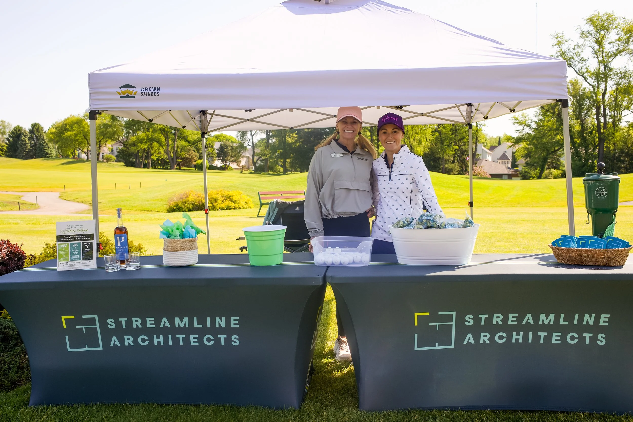 Two women standing under a white canopy tent at an outdoor event on a sunny day, smiling at the camera. The table in front of them is covered with a black tablecloth displaying the logo and name of Streamline Architects. The table has various items i