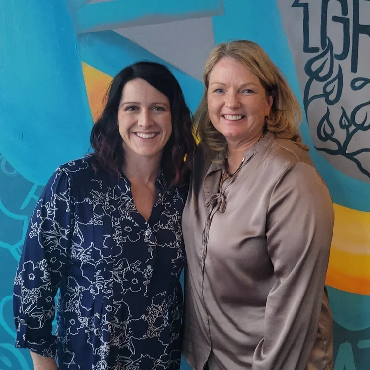 Two women smiling and standing side by side in front of a colorful mural background.