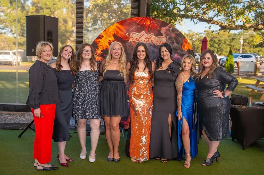 A group of eight women dressed in formal attire, standing together outdoors at night in front of a decorated backdrop with flames and the text "Fuel the Fire".