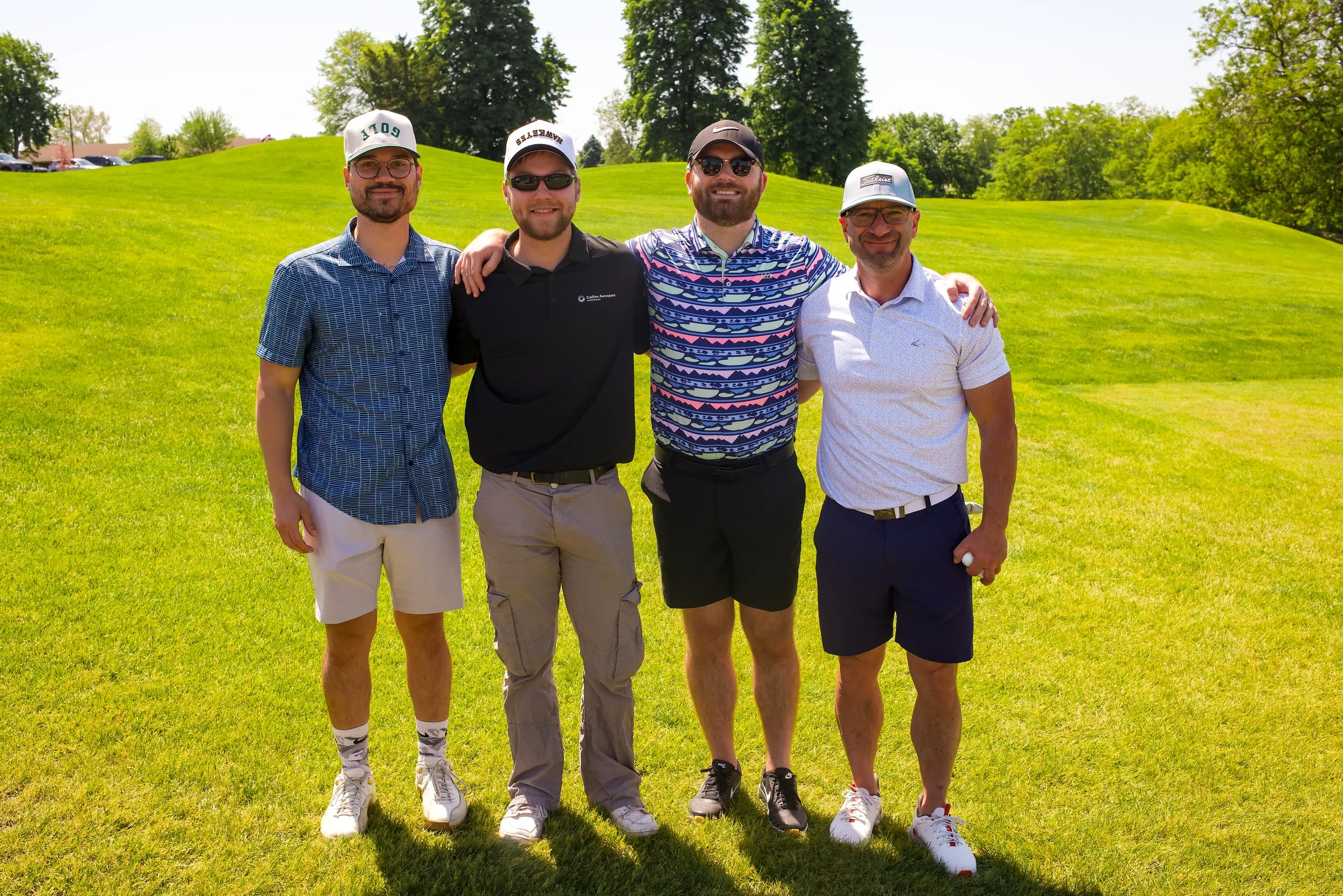 Four men standing on a grassy golf course, smiling and posing for a photo on a sunny day, with trees in the background.