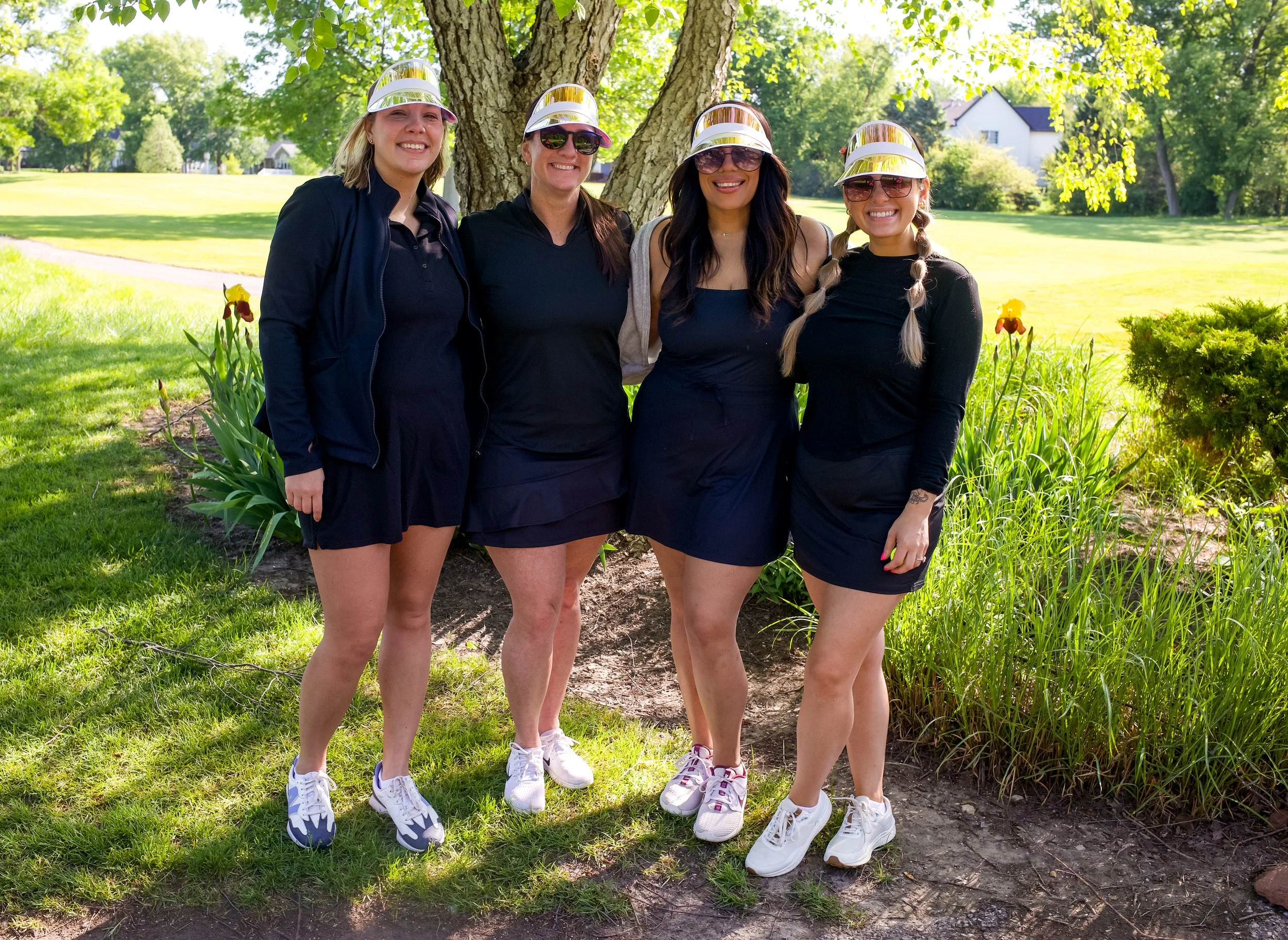 Four women standing outdoors near a tree, smiling and wearing protective face shields, sunglasses, and casual black outfits.