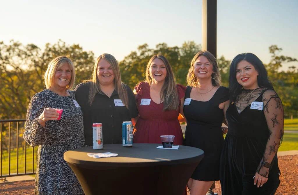 Five women standing outdoors around a high table at sunset, smiling and holding drinks, with a park in the background.