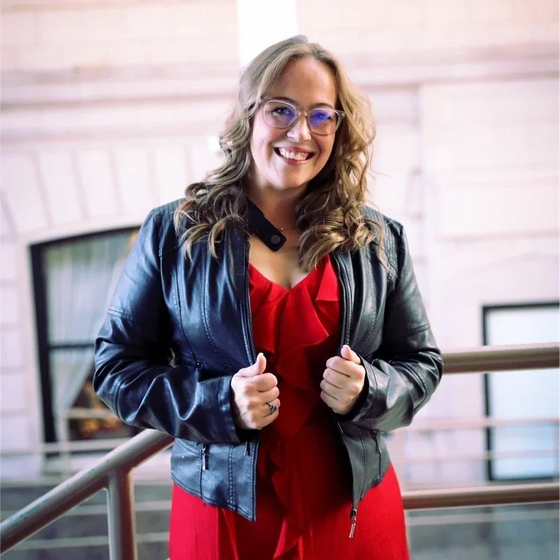 A woman with wavy blonde hair and glasses smiling, wearing a black leather jacket over a red dress, standing next to a railing indoors.