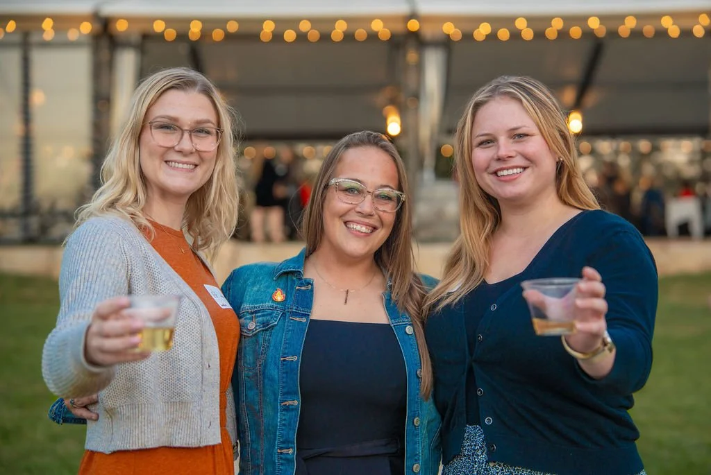 Three smiling women standing outdoors at an event, holding small cups, with string lights above them.