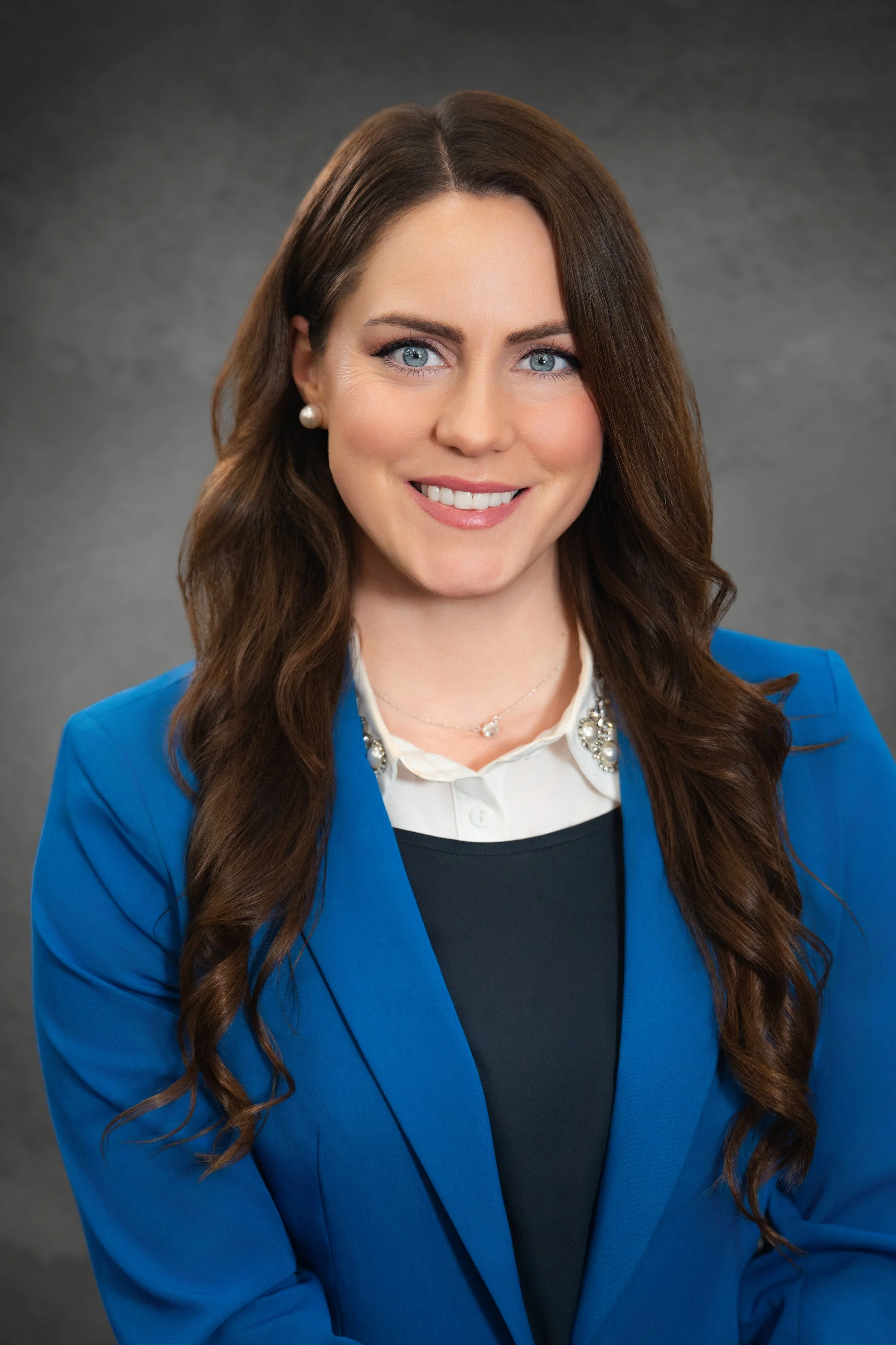 Professional woman with long wavy brown hair and blue eyes, wearing a blue blazer, white blouse, pearl earrings, and necklace against a dark gray background.