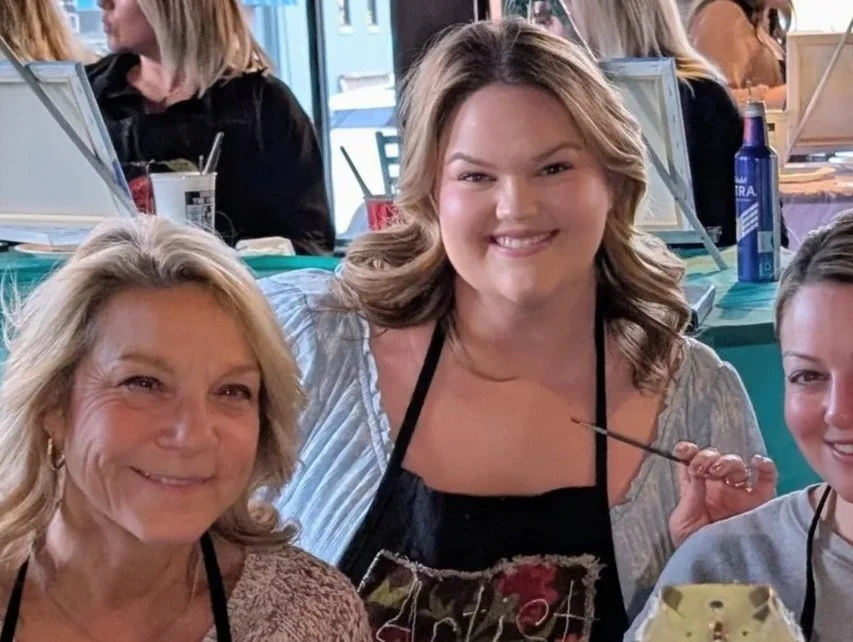 Three women smiling at a restaurant or cafe, with paintings supplies and beverages on the table.