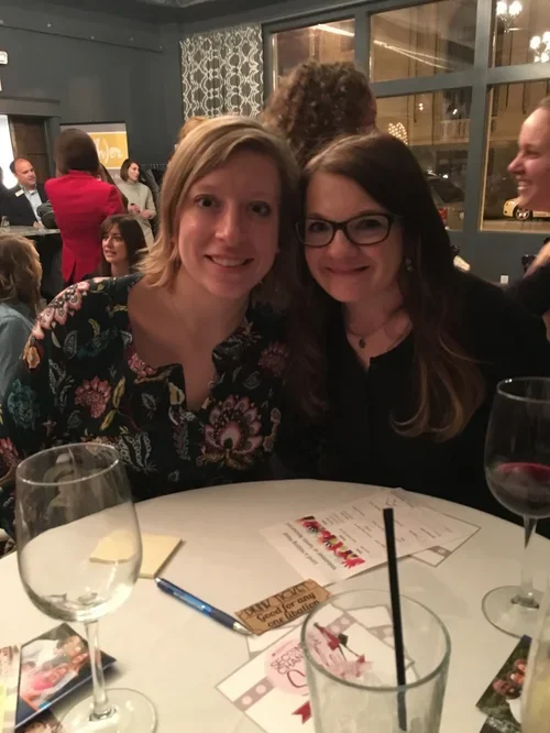 Two women sitting at a table in a restaurant or bar, smiling at the camera. The table has glasses and some paper menus or programs on it. Other people are seen in the background, socializing near large windows.