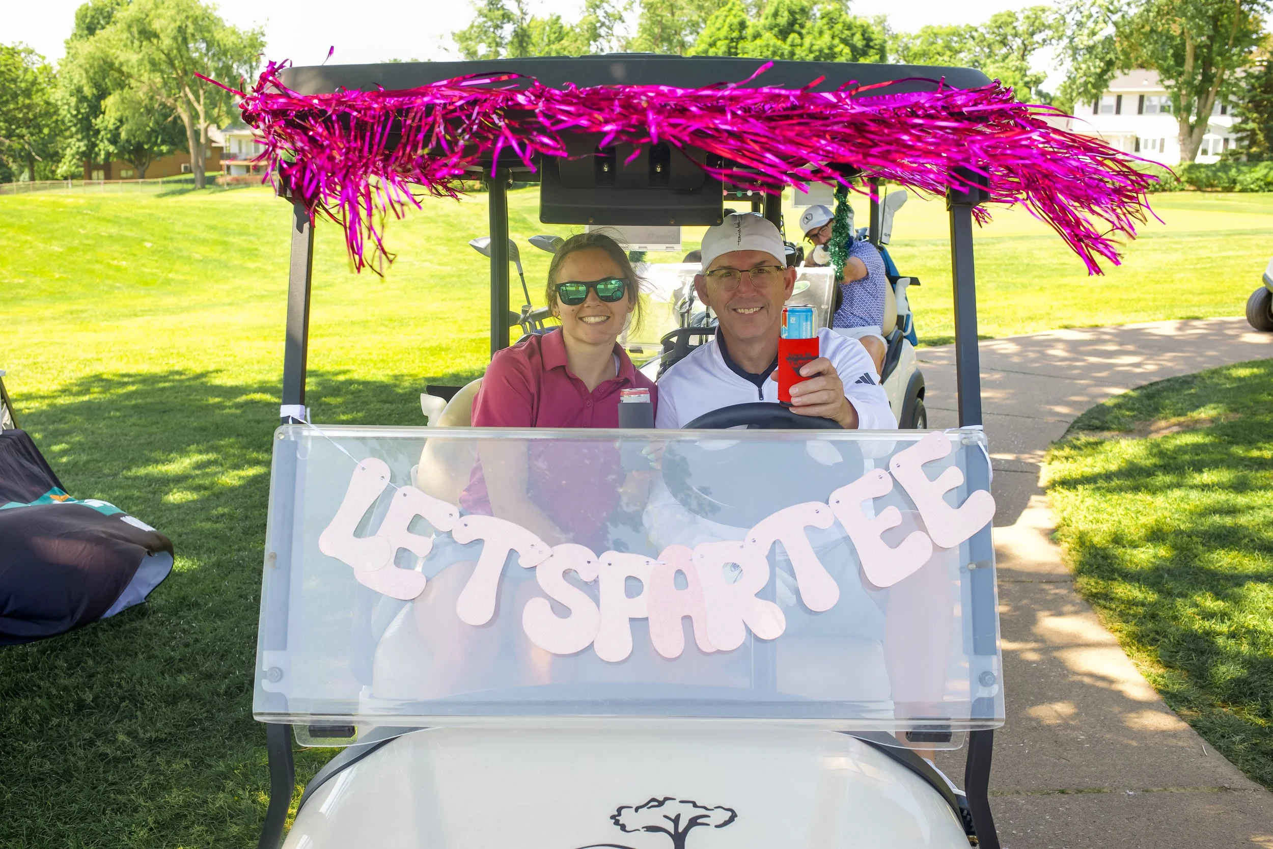 A golf cart decorated for a celebration, with a pink tinsel garland on the top and a white paper banner that says "Happy Eather" hung across the front. Two people sit inside the cart, smiling and holding drinks, while a person in the back adjusts som