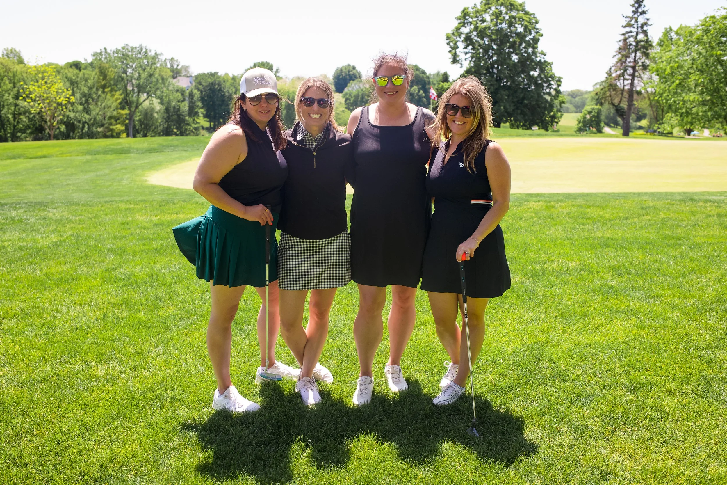 Four women standing on a golf course, smiling, with trees and grass in the background, holding golf clubs.