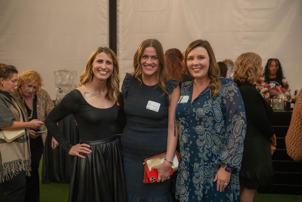 Three women smiling at an indoor event, standing close together with other attendees in the background.