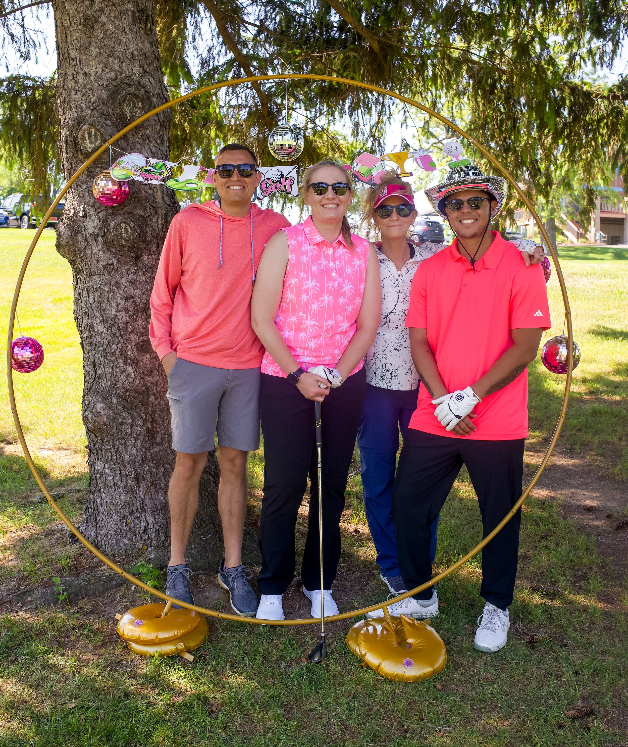 Group of four people standing outdoors behind a large inflatable gold hoop, with a tree in the background. They are smiling and wearing sporty clothing, with some wearing sunglasses and a hat. There are colorful decorations with golf-related designs 