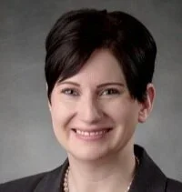 A headshot of a woman with short dark hair, smiling, wearing a black blazer and pearl necklace against a gray background.