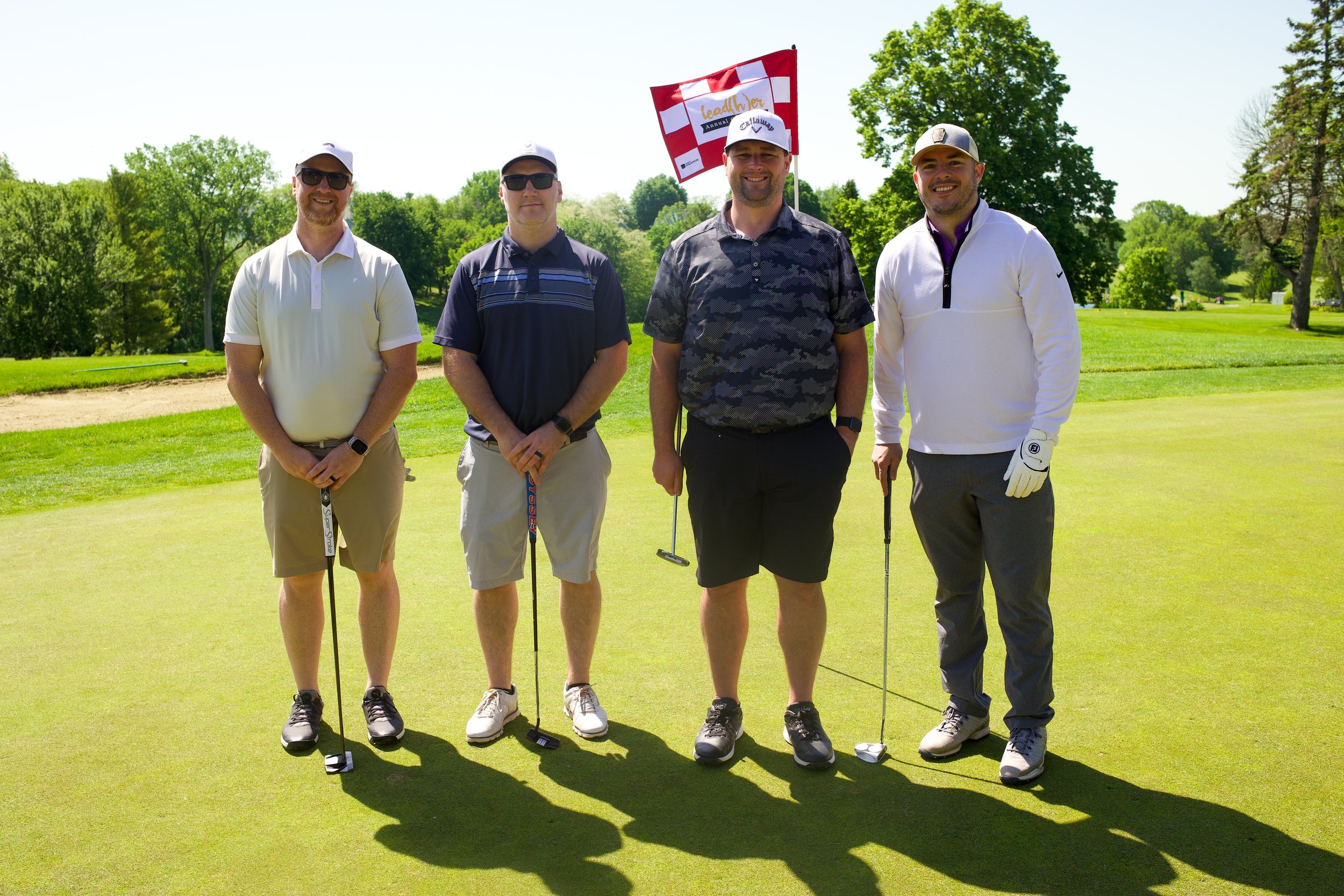 Four men standing on a golf course, holding golf clubs, smiling, dressed in golf attire, with trees and a blue sky in the background.