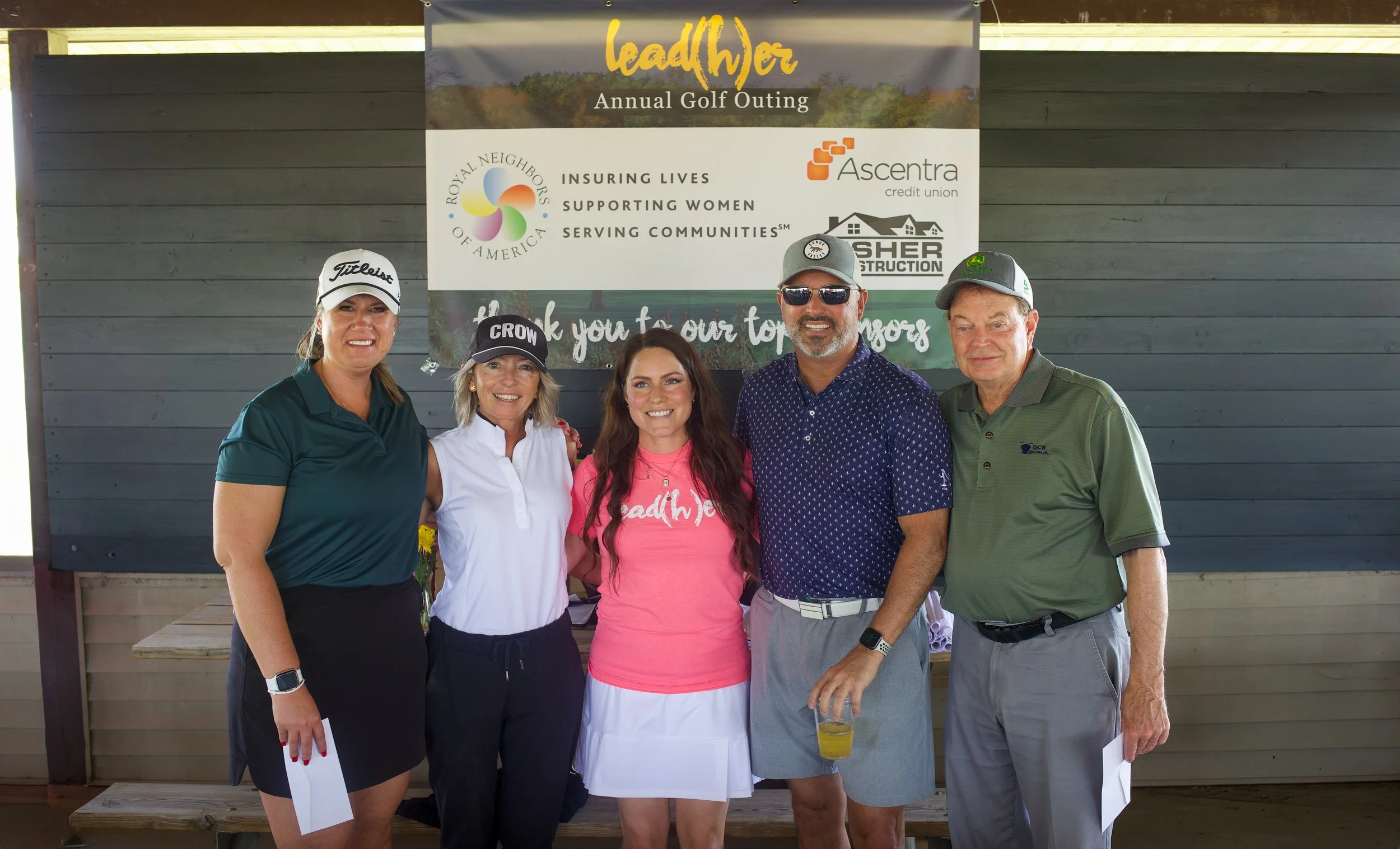 A group of five people standing together at an outdoor event, smiling for the camera. Behind them is a banner that reads "lead(h)ers Annual Golf Outing" and includes logos of various sponsors. The people are dressed in casual and golf attire.