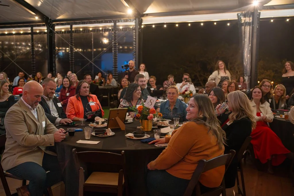A group of people seated at round tables at a formal event, some smiling and engaging, with string lights and dark outdoor scenery in the background.