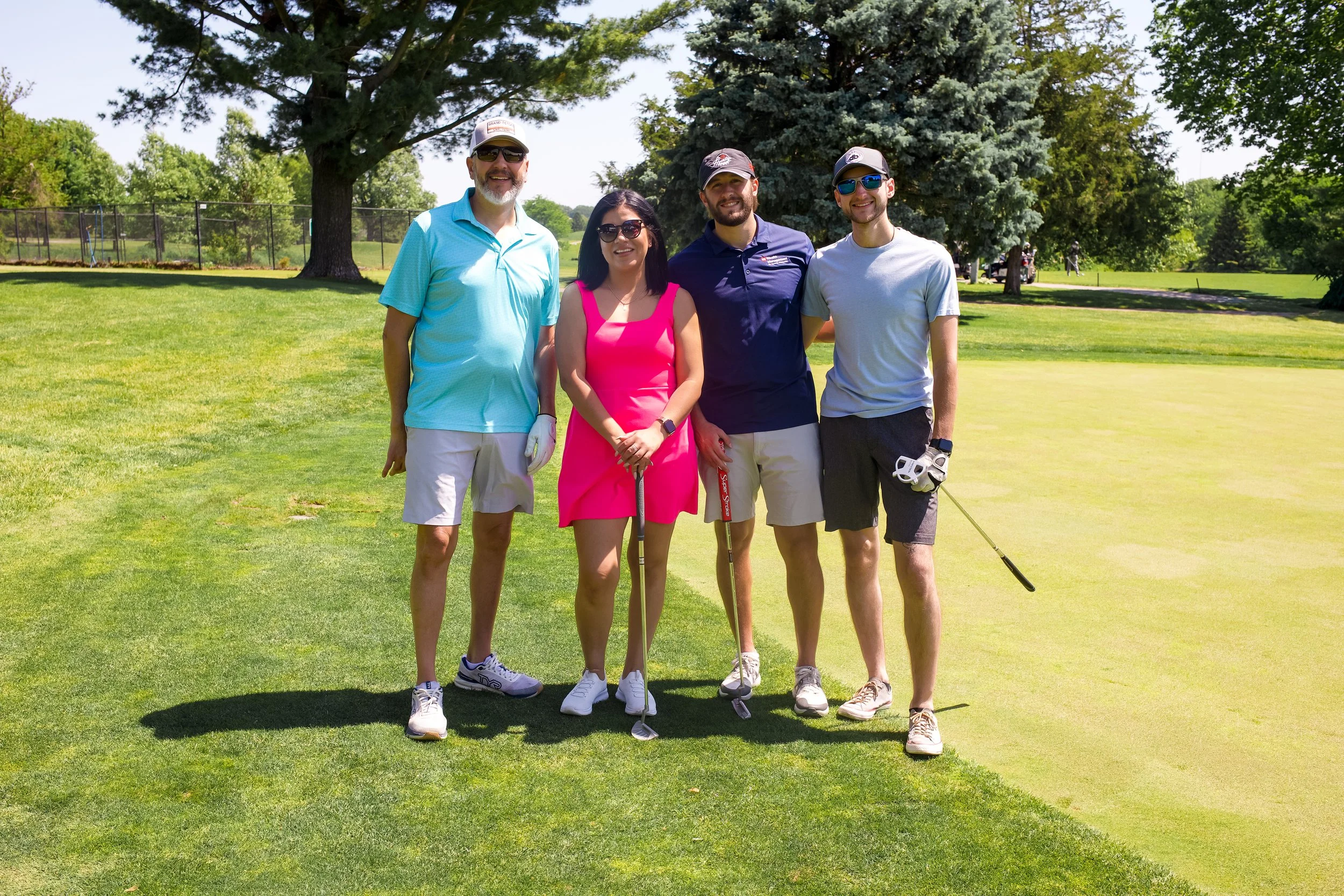 Group of five people standing on a golf course with trees in the background, smiling for the camera, holding golf clubs.
