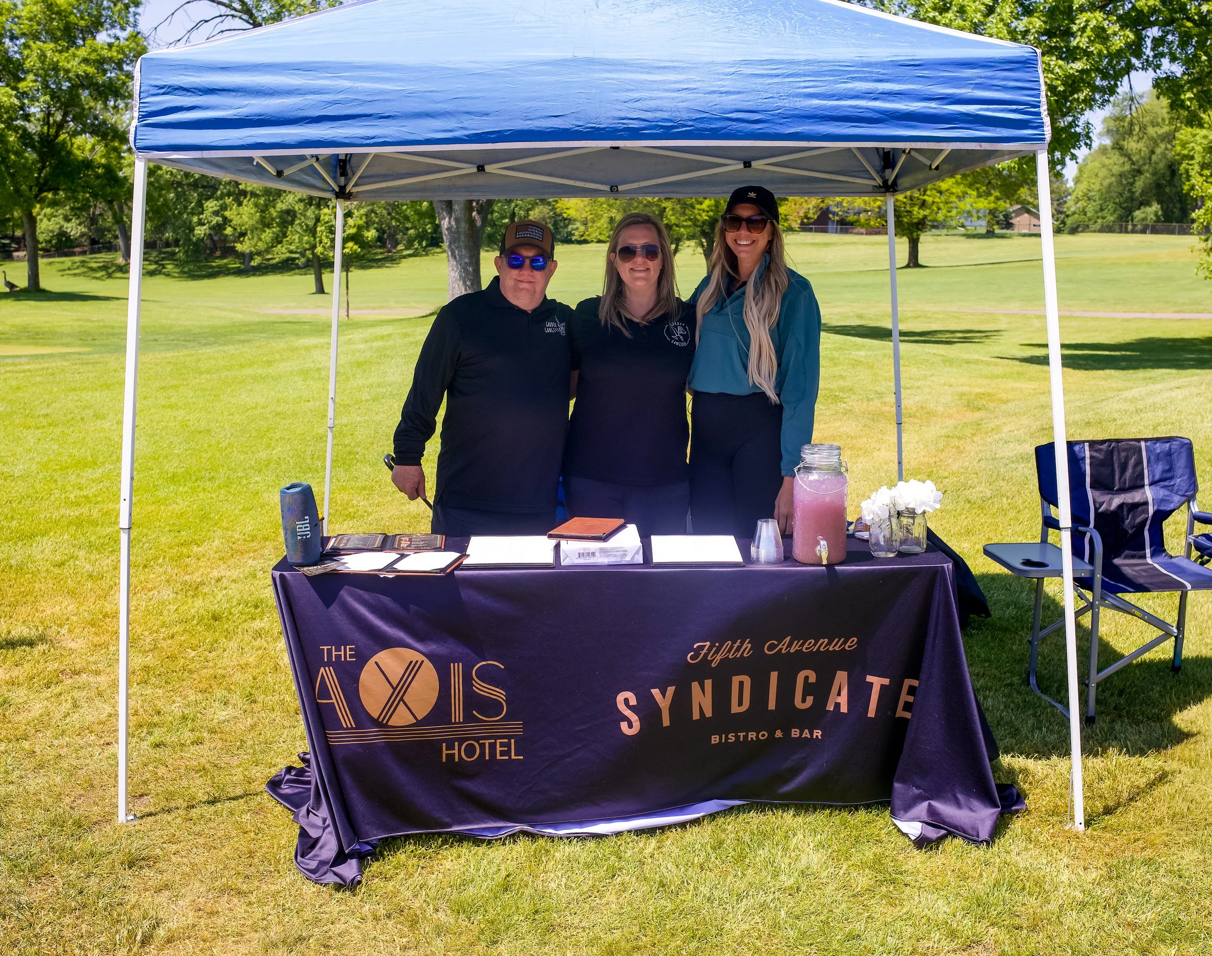 Three people standing under a blue pop-up tent at an outdoor event, smiling. Two women and one man, all wearing sunglasses, are behind a table with a dark purple tablecloth displaying the logo and name of a hotel and bar, with a park and trees in the