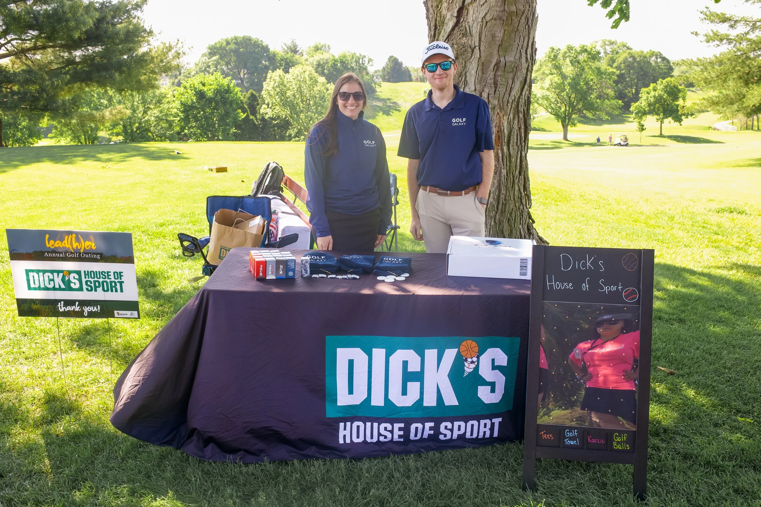 Two people, a man and a woman, smiling and standing behind a table at an outdoor event on a golf course. The table displays various golf-related items and signs for a charity event called 'Dick's House of Sport,' with lush green trees and sunny sky i