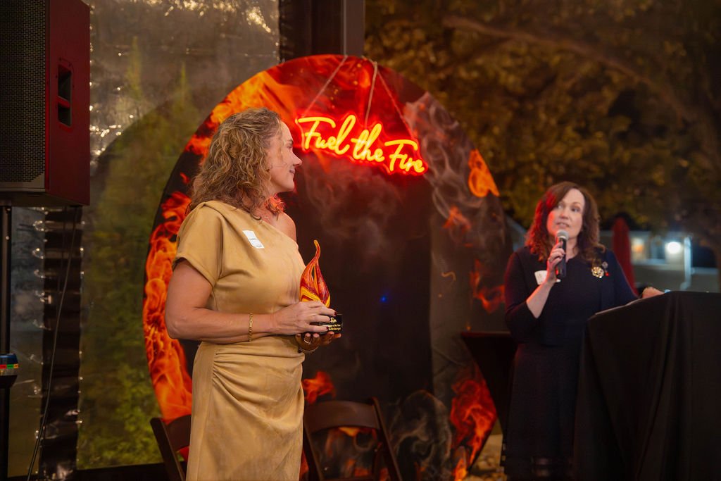 Two women at an event, one holding an award or trophy, with a backdrop of a neon sign saying 'Fuel the Fire' and a fiery design.