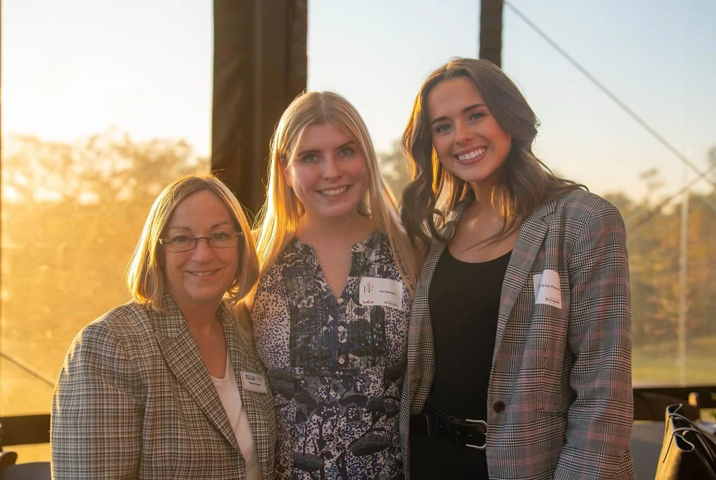 Three women smiling outdoors during sunset, wearing business casual attire, standing close together.