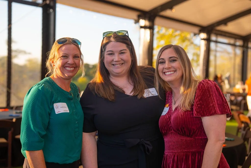 Three women smiling and standing close together at an outdoor event, wearing casual and semi-formal clothing, under a canopy with trees and other people in the background.