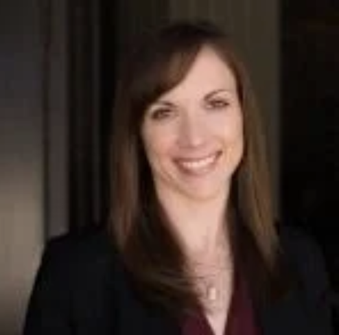 A woman with shoulder-length brown hair, smiling, wearing a black blazer and a maroon top, standing indoors.