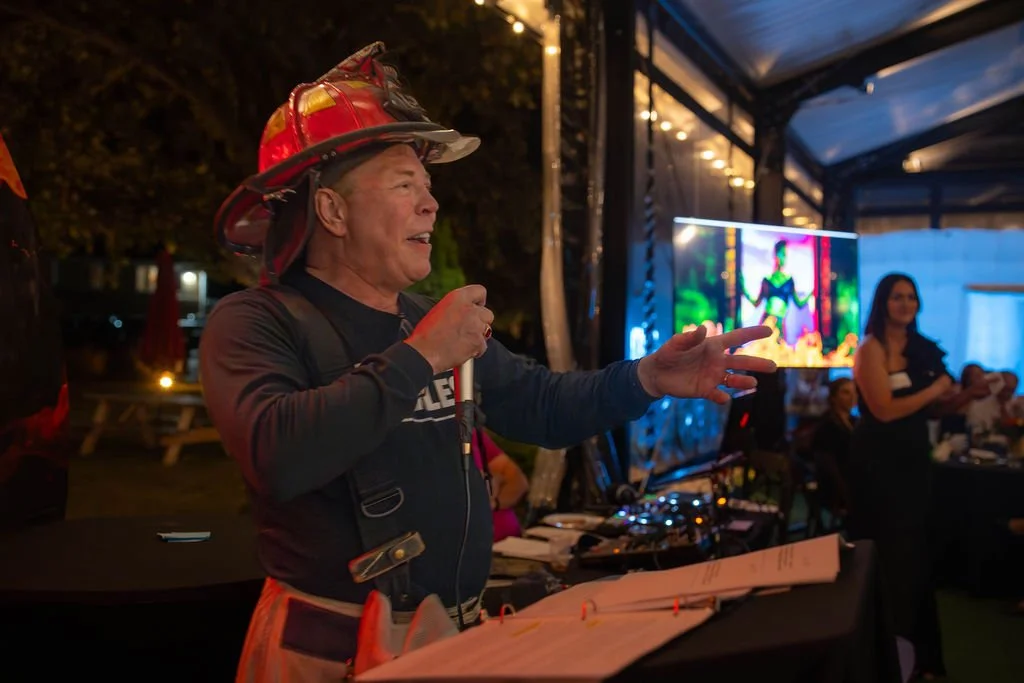 A man wearing a firefighter helmet and a dark shirt is speaking into a microphone at an indoor event with a screen and people seated in the background.