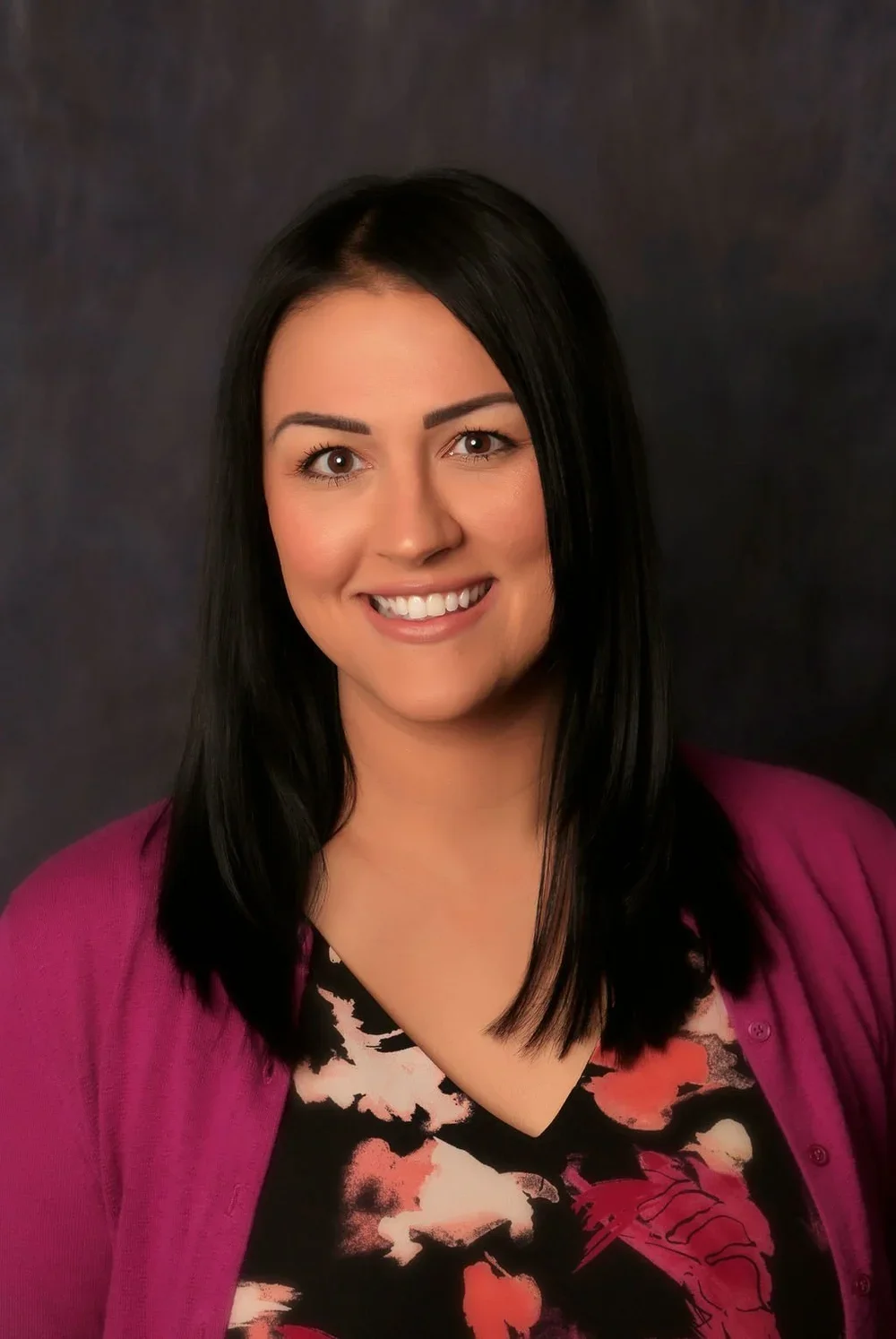 Professional headshot of a woman with dark straight hair, smiling, wearing a purple cardigan over a floral blouse, against a dark background.