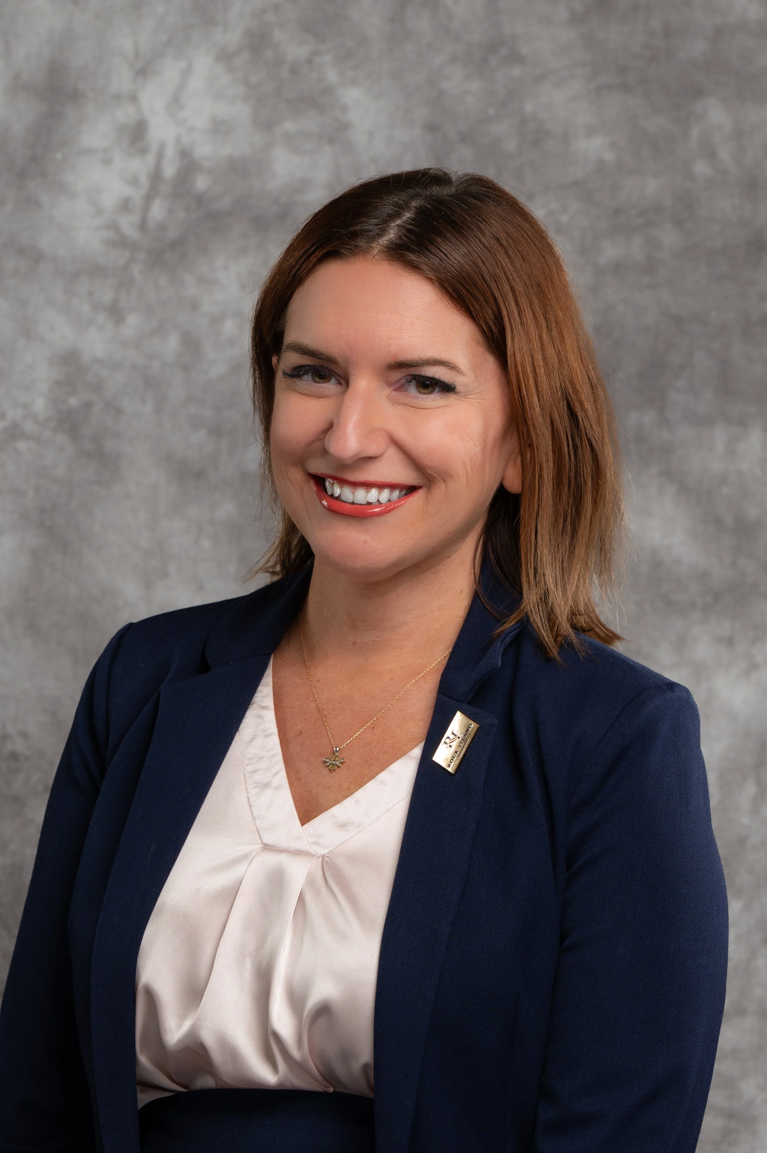 Professional woman with brown hair and a friendly smile, wearing a navy blazer and a white blouse, standing against a gray background.