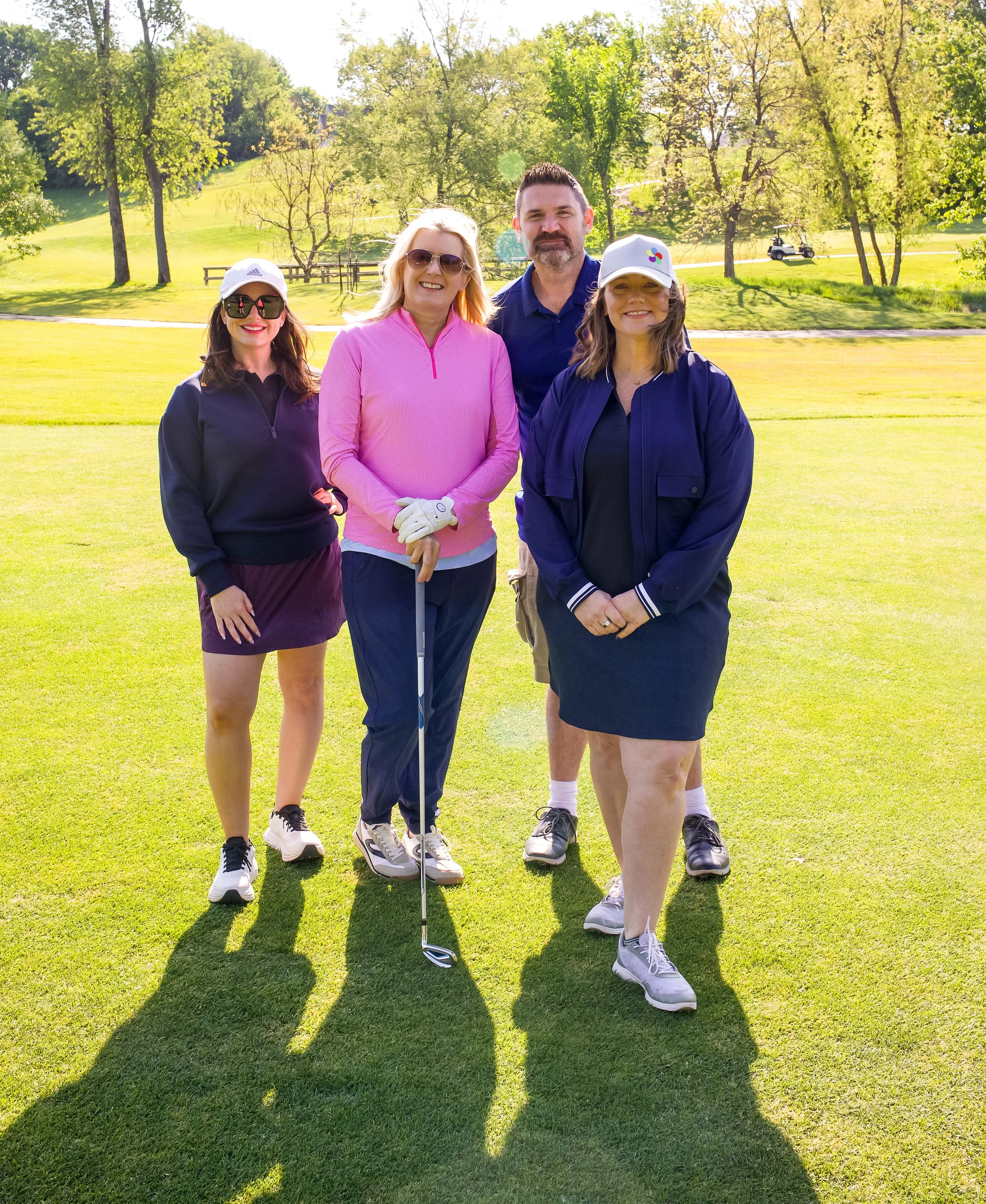 Group of four people standing on a golf course, with trees and a golf cart in the background. Three women and one man, dressed in golf attire, smiling and posing for the photo, with their shadows cast on the grass.